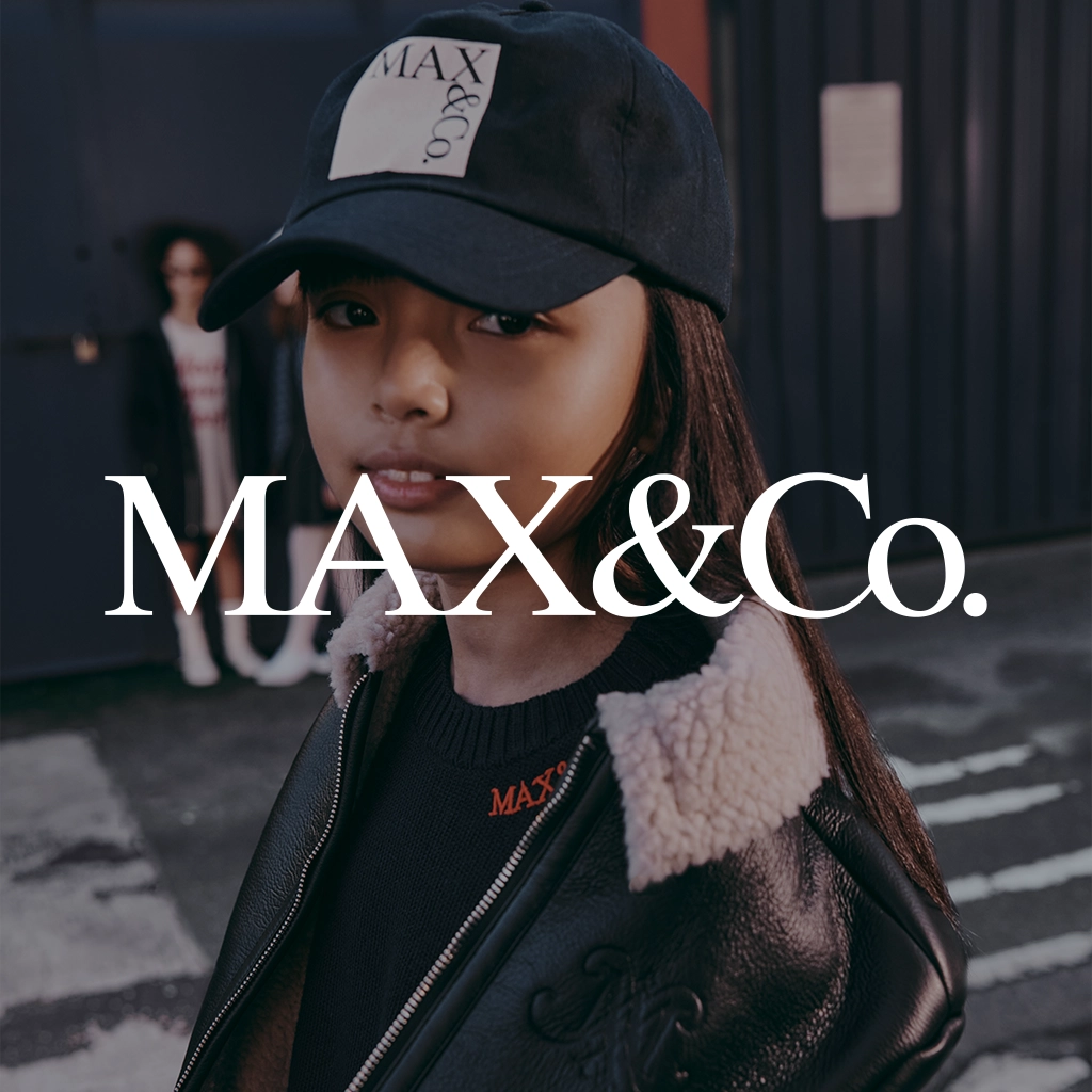 A child model wearing a black MAX&Co. baseball cap, a black leather jacket with a cream shearling collar, and a black knit top with an orange logo, set against a dark urban street background with a large white MAX&Co.